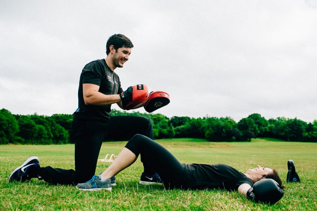 Personal trainer holding pads while client performs boxing sit-ups during an outdoor fitness session.