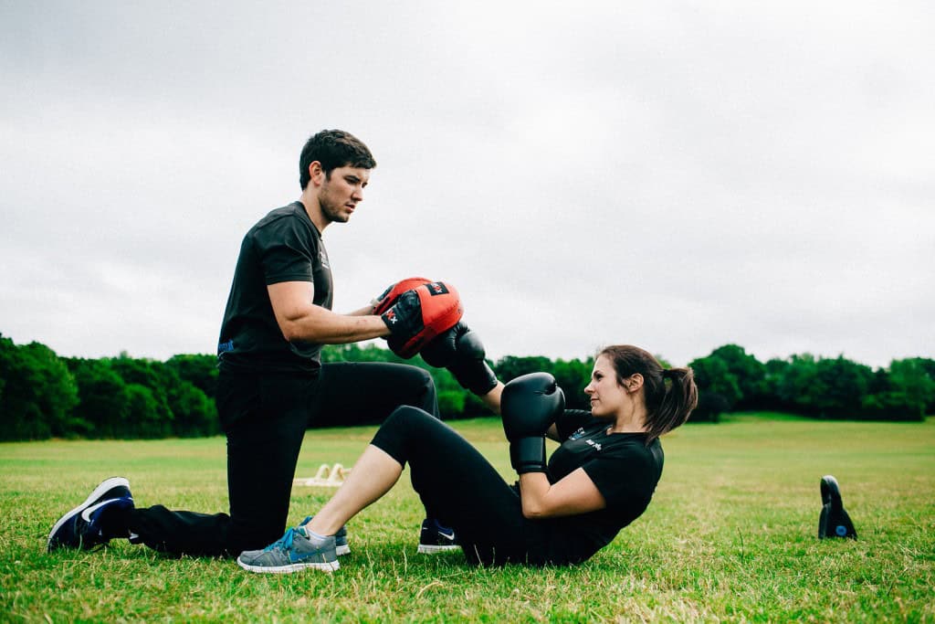 Female client doing sit-ups with boxing gloves while trainer holds pads during an outdoor personal training session in Essex park